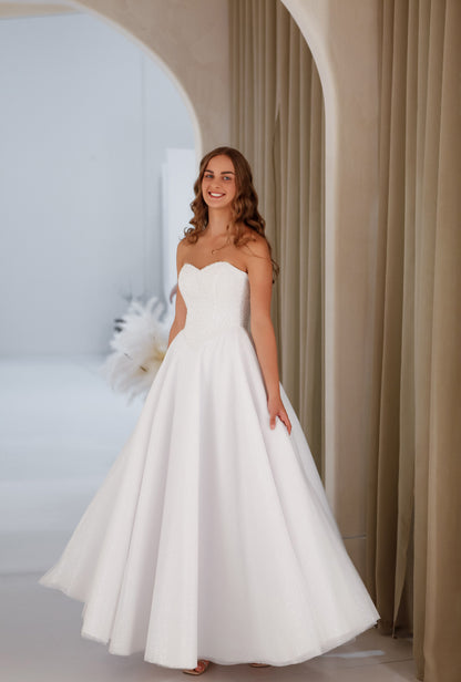 Woman in a white strapless gown standing in a softly lit room with neutral decor.