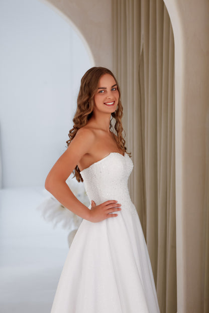 Woman in a white strapless gown standing in a softly lit room with neutral walls.