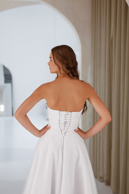 Woman wearing a white strapless dress with a lace-up back in a neutral setting.