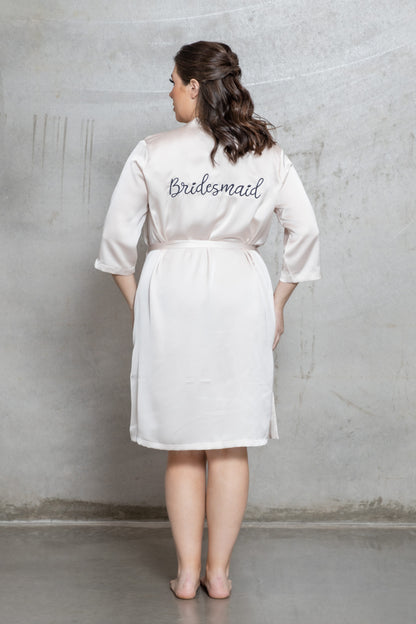 Woman wearing a white satin robe with 'Bridesmaid' embroidery against a gray concrete wall
