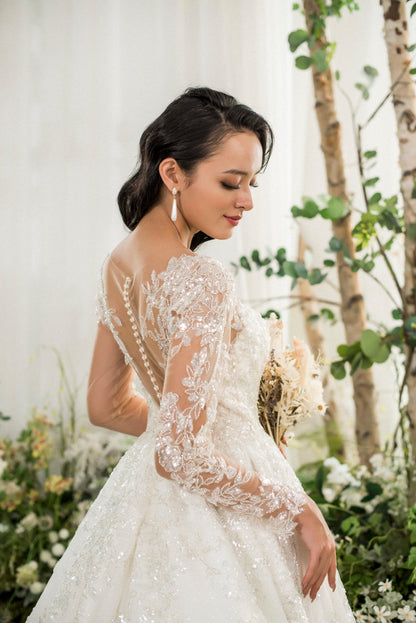 Woman in a white wedding dress with intricate lace details standing in a natural setting with trees and flowers
