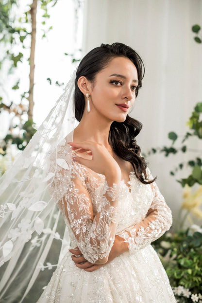 Woman in a white wedding dress with a veil, standing outdoors with greenery in the background