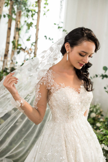 Woman in a white wedding dress with a veil, standing outdoors with greenery in the background