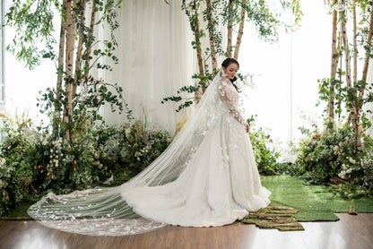Woman in a white wedding dress with a long train standing in a decorated indoor setting with flowers and greenery