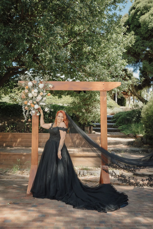 Woman in a black dress with a long train standing in front of a wooden archway with floral decorations, surrounded by trees.
