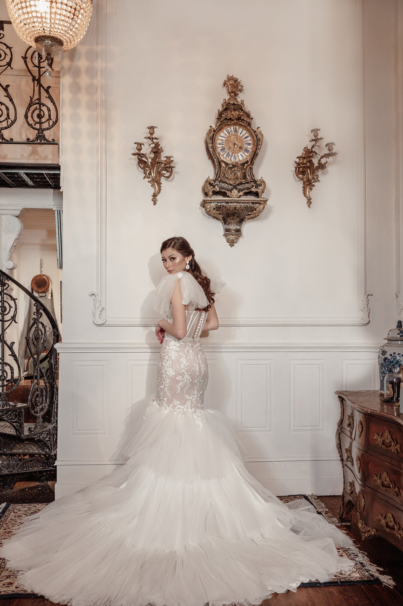 Woman in a white wedding dress standing in an elegant room with decorative clock and furniture.