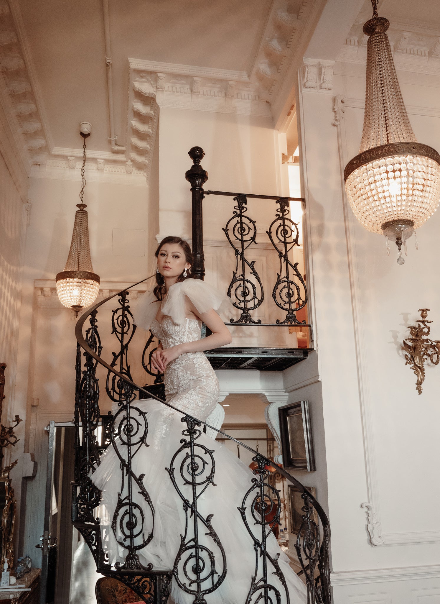 Woman in a white dress standing on a decorative staircase with chandeliers above.