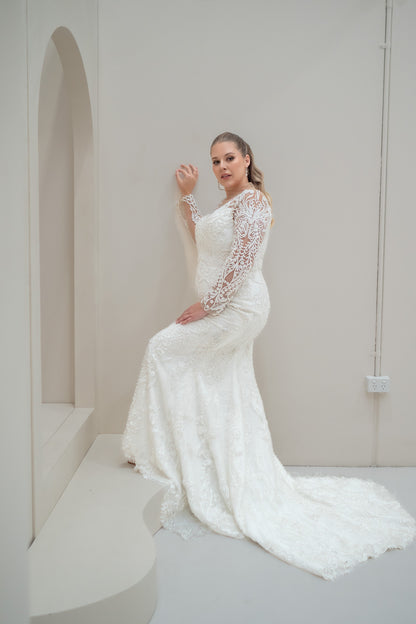 Woman in a white lace wedding dress sitting on a white bench against a plain white wall.