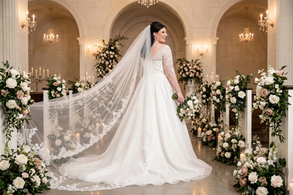 A bride in a white lace wedding gown and long sheer veil stands in an arched hall decorated with floral arrangements and candles, holding a bouquet.