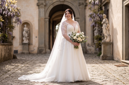 A bride in a white lace wedding dress with a long veil holds a bouquet of white and cream flowers, standing in a sunlit stone courtyard with arches and statues in the background.