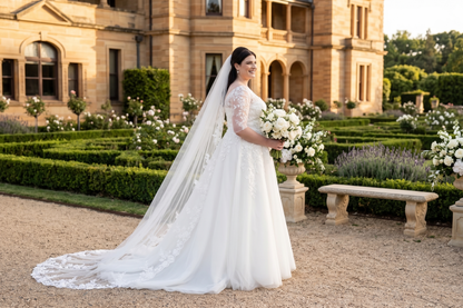 A bride in a white lace wedding dress with a long sheer veil holds a bouquet of white flowers, standing in a formal garden courtyard with stone buildings and hedges in the background.