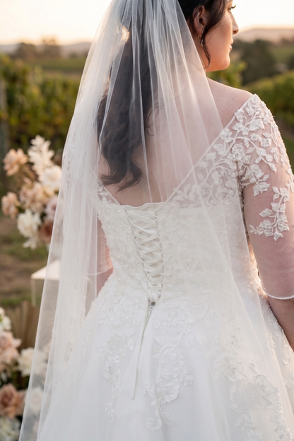A bride shown from the back wearing a white lace wedding dress with a long sheer veil, photographed outdoors in warm natural light.