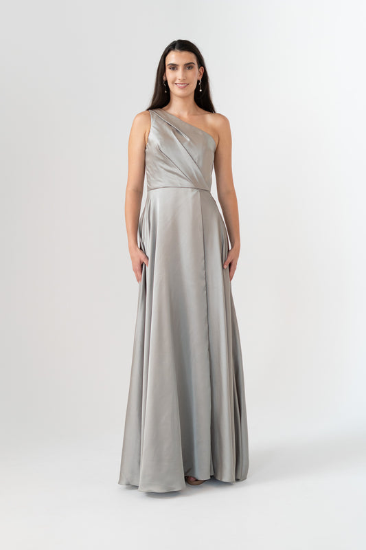 A woman modeling a floor-length, one-shoulder formal gown in a light silver-gray satin fabric against a plain light gray studio background.