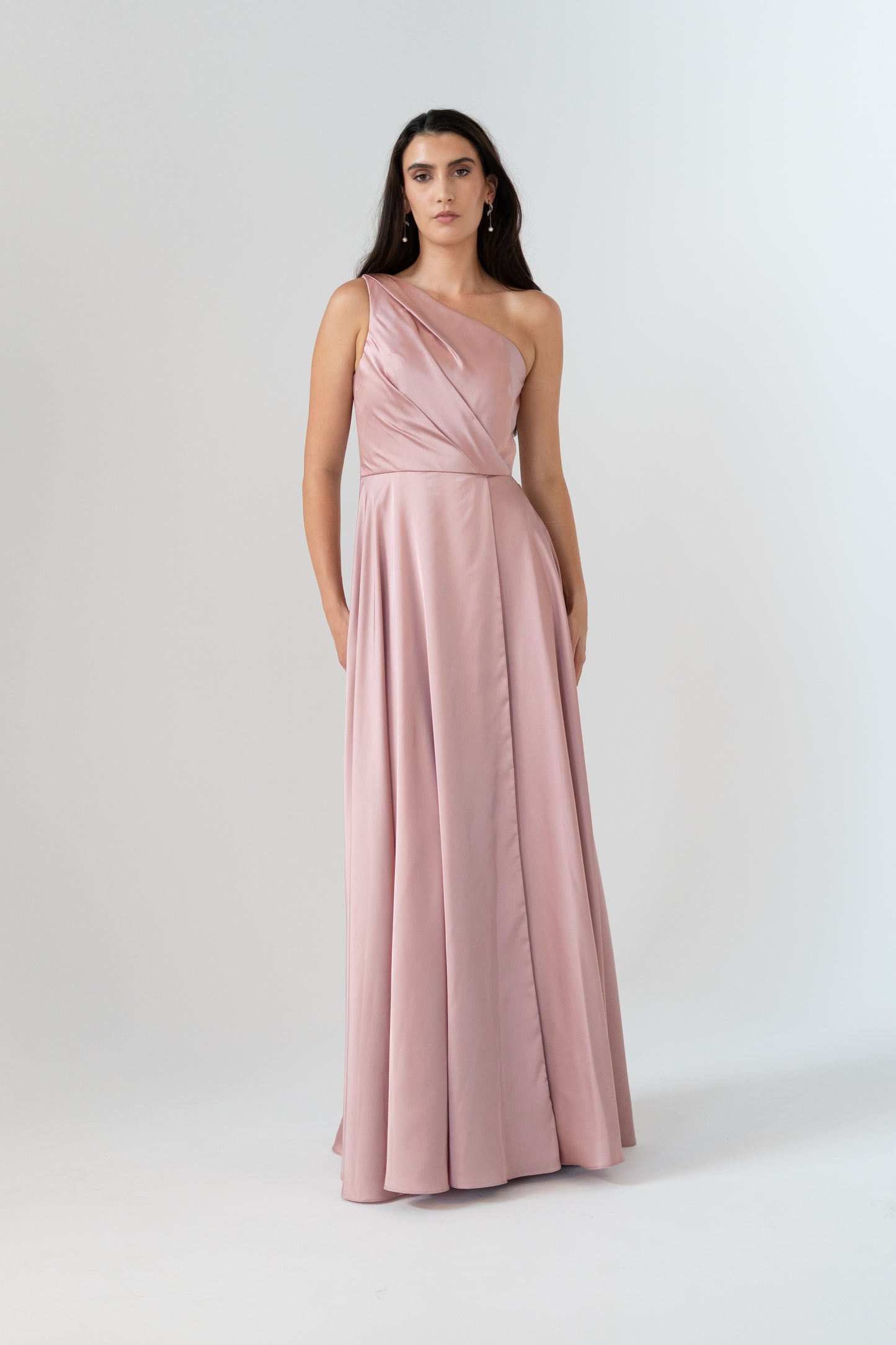 A woman modeling a floor-length, one-shoulder dusty pink gown with a draped bodice and flowing skirt, photographed against a plain light gray studio background.