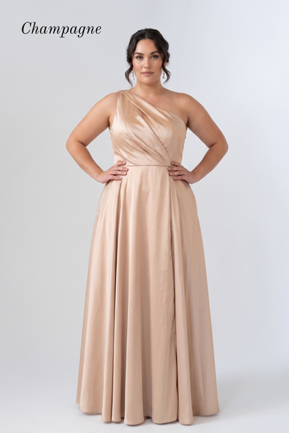 A woman modeling a floor-length champagne one-shoulder gown against a light gray studio background. The word 'Champagne' appears in the upper left corner.