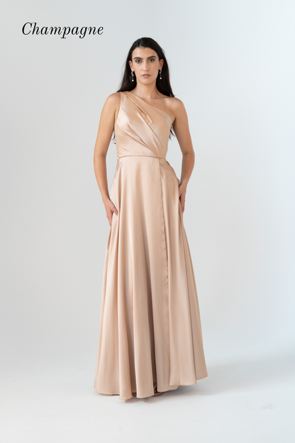 A woman modeling a floor-length champagne satin gown with a draped one-shoulder neckline and a flowing skirt, standing against a light gray studio background. The word 'Champagne' appears in the upper left corner.