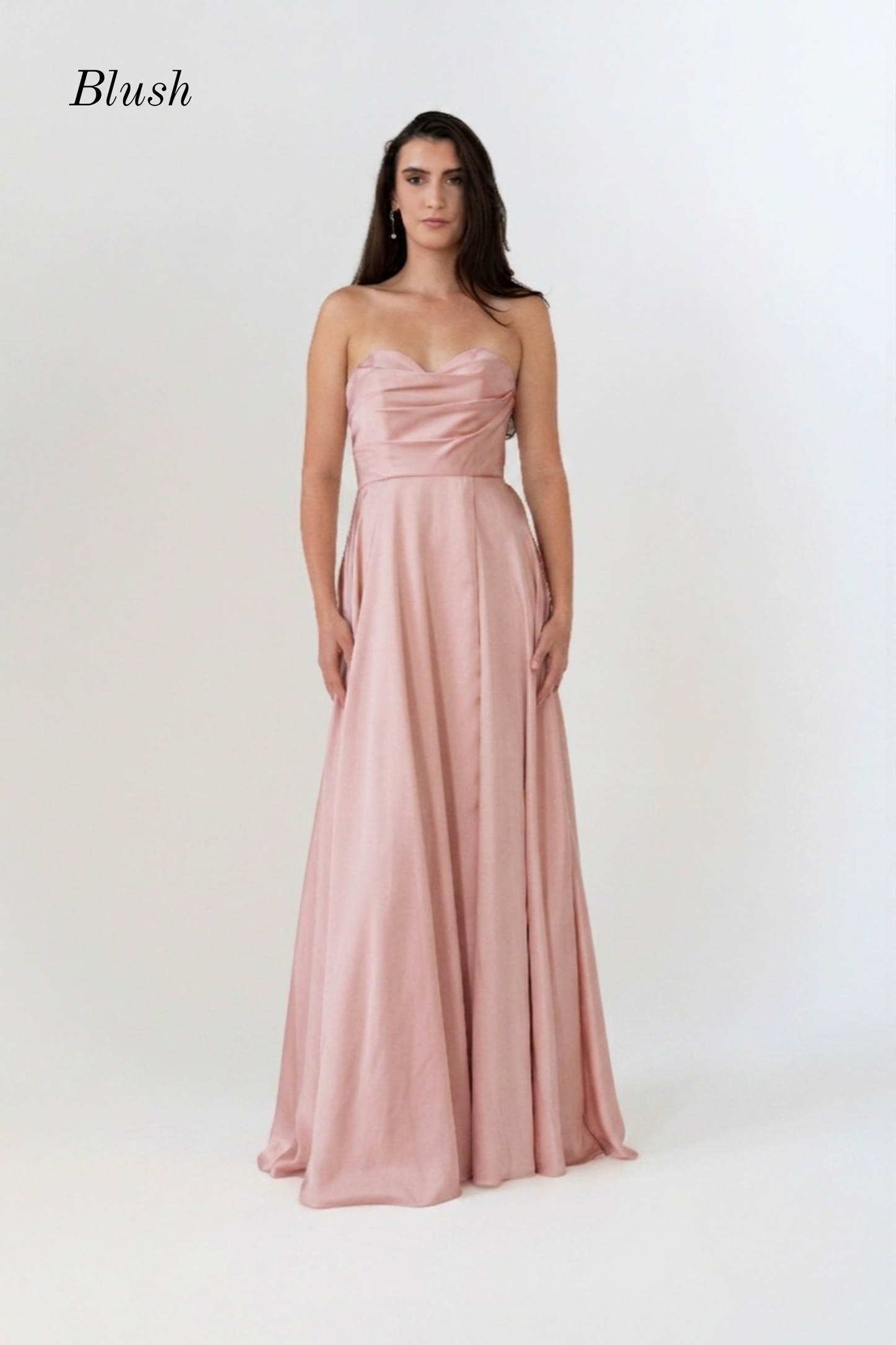 A woman modeling a floor-length blush pink gown with a sweetheart neckline, fitted bodice, and flowing skirt against a plain light background. The word 'Blush' appears in the upper left corner.