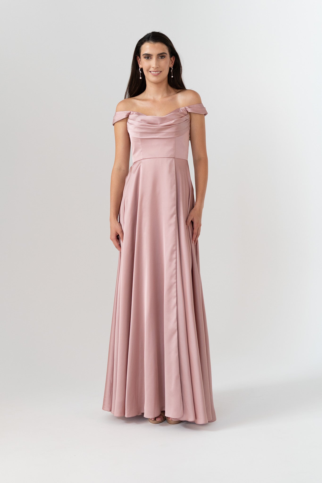 A woman modeling a floor-length off-the-shoulder gown in dusty pink satin, standing against a light gray studio background.