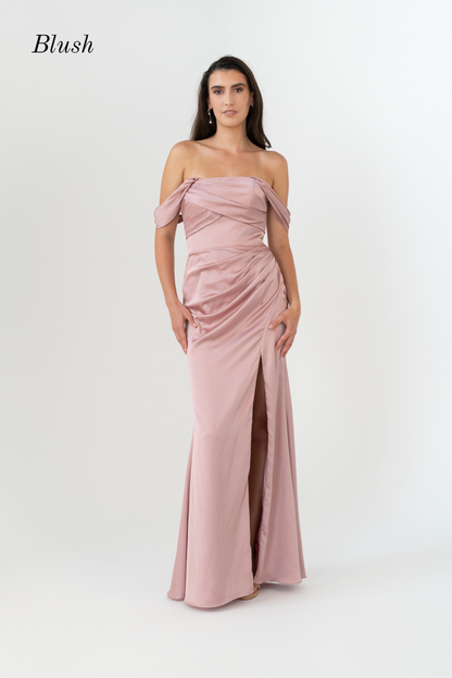 A woman modeling a blush pink off-the-shoulder floor-length gown with ruched draping and a front slit, photographed against a clean light gray studio background. The word 'Blush' appears in the upper left corner.