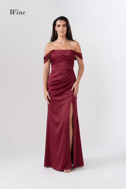A woman modeling an off-the-shoulder floor-length gown in a deep wine red satin fabric, featuring a ruched bodice and a high front slit, against a plain light gray studio background.
