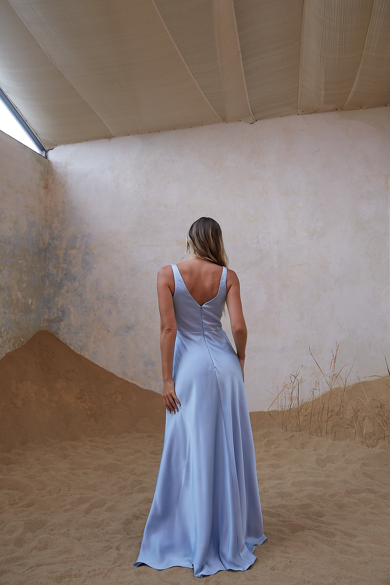 A woman stands with her back to the camera wearing a light blue sleeveless maxi dress with a deep V-back and a flowing satin-like skirt, posed in a studio set with textured beige walls and sand-like floor props.