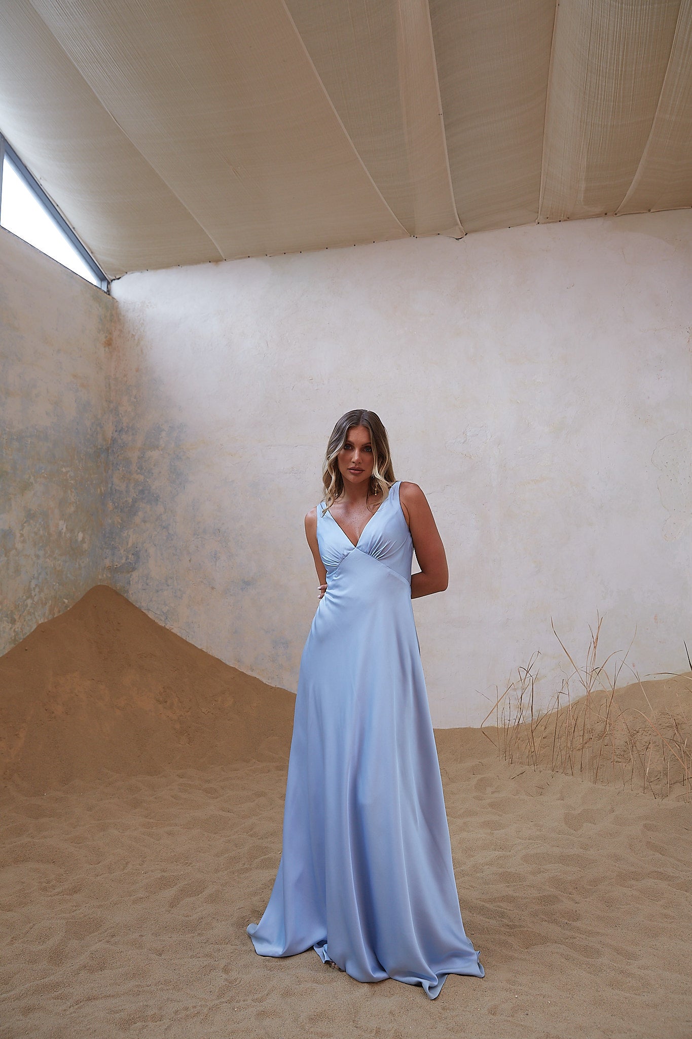 Woman in a light blue dress standing in a sandy, sunlit room with a textured wall.