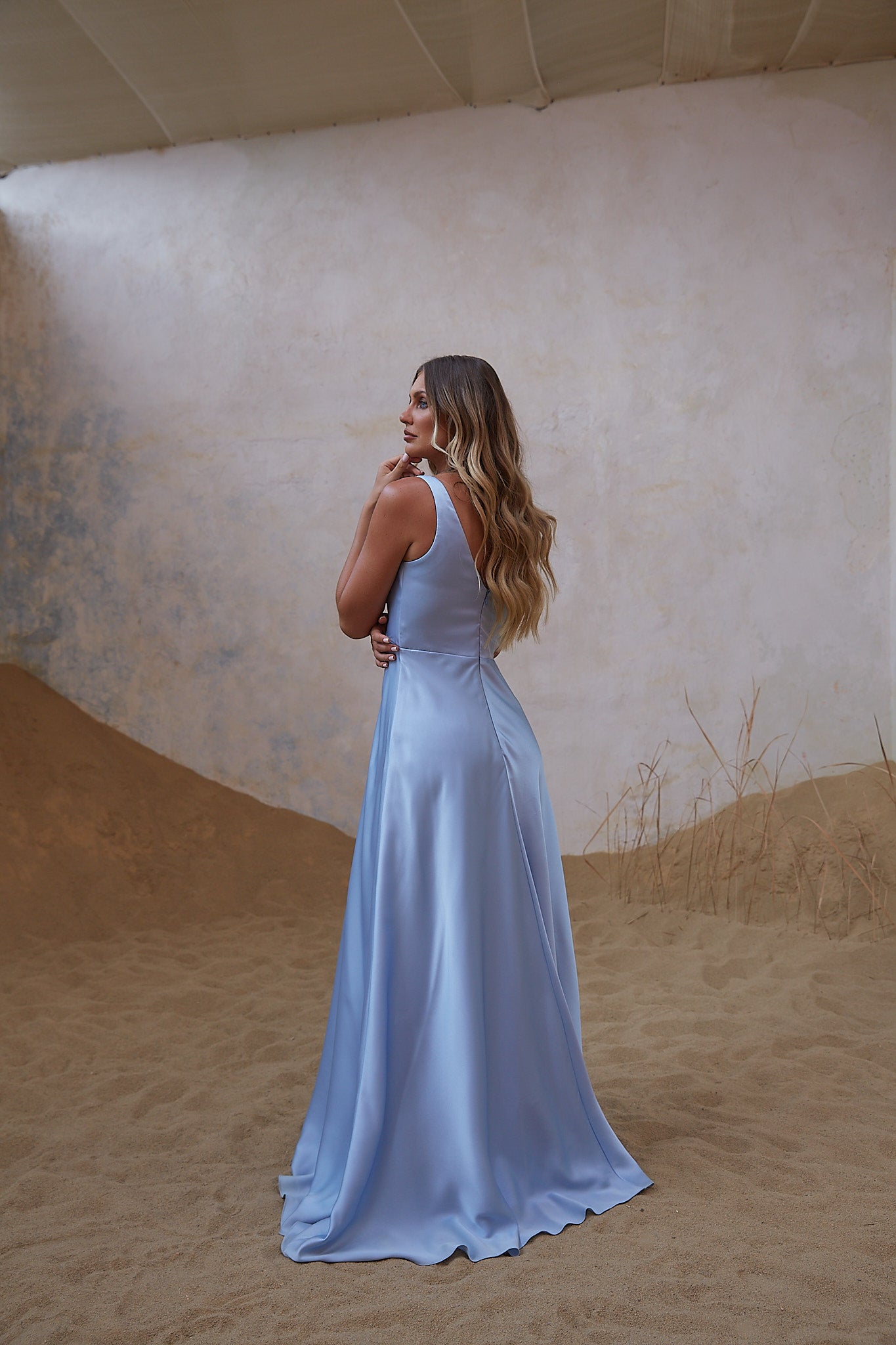 A woman wearing a sleeveless light blue maxi dress stands in profile against a textured beige wall and sandy floor, with a draped fabric backdrop behind her.