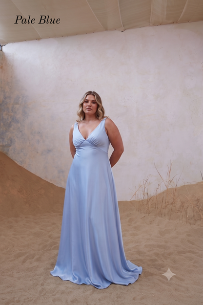 A woman modeling a sleeveless pale blue floor-length gown with a deep V-neckline, standing in a sandy studio setup with a textured beige backdrop. The words 'Pale Blue' appear in the upper left corner.