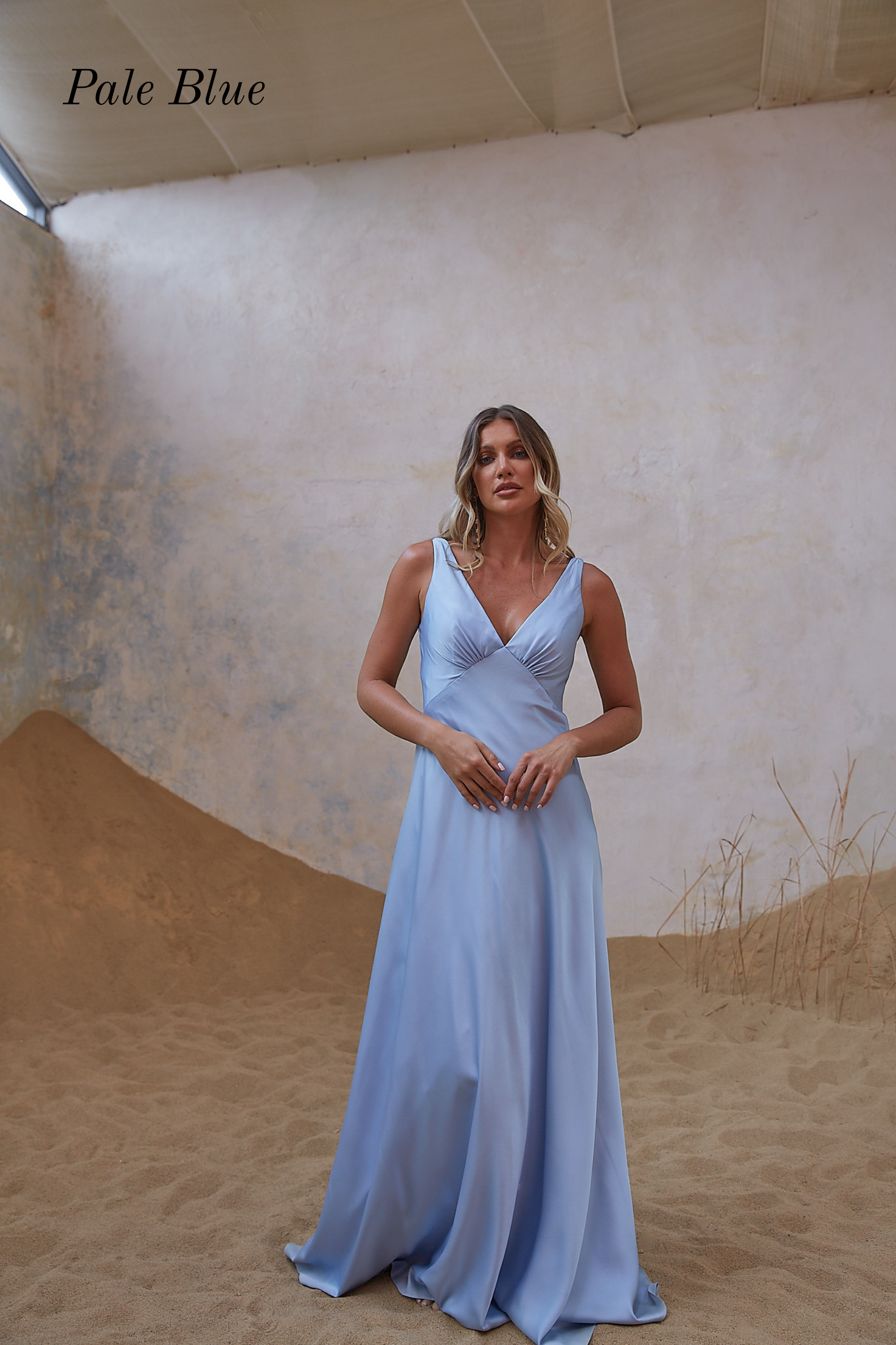 A woman modeling a sleeveless pale blue floor-length gown in a sandy indoor set with textured walls. The text 'Pale Blue' appears in the upper left corner.