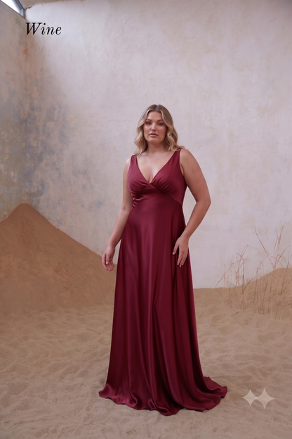 A woman modeling a floor-length wine red satin gown with a deep V-neckline and sleeveless design, set against a textured beige backdrop with sand dunes.