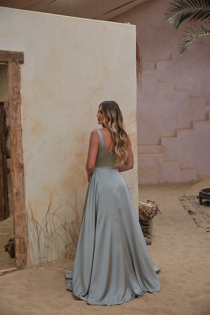 Woman in a long, light-colored dress standing in a desert-like setting with sand and minimal vegetation.