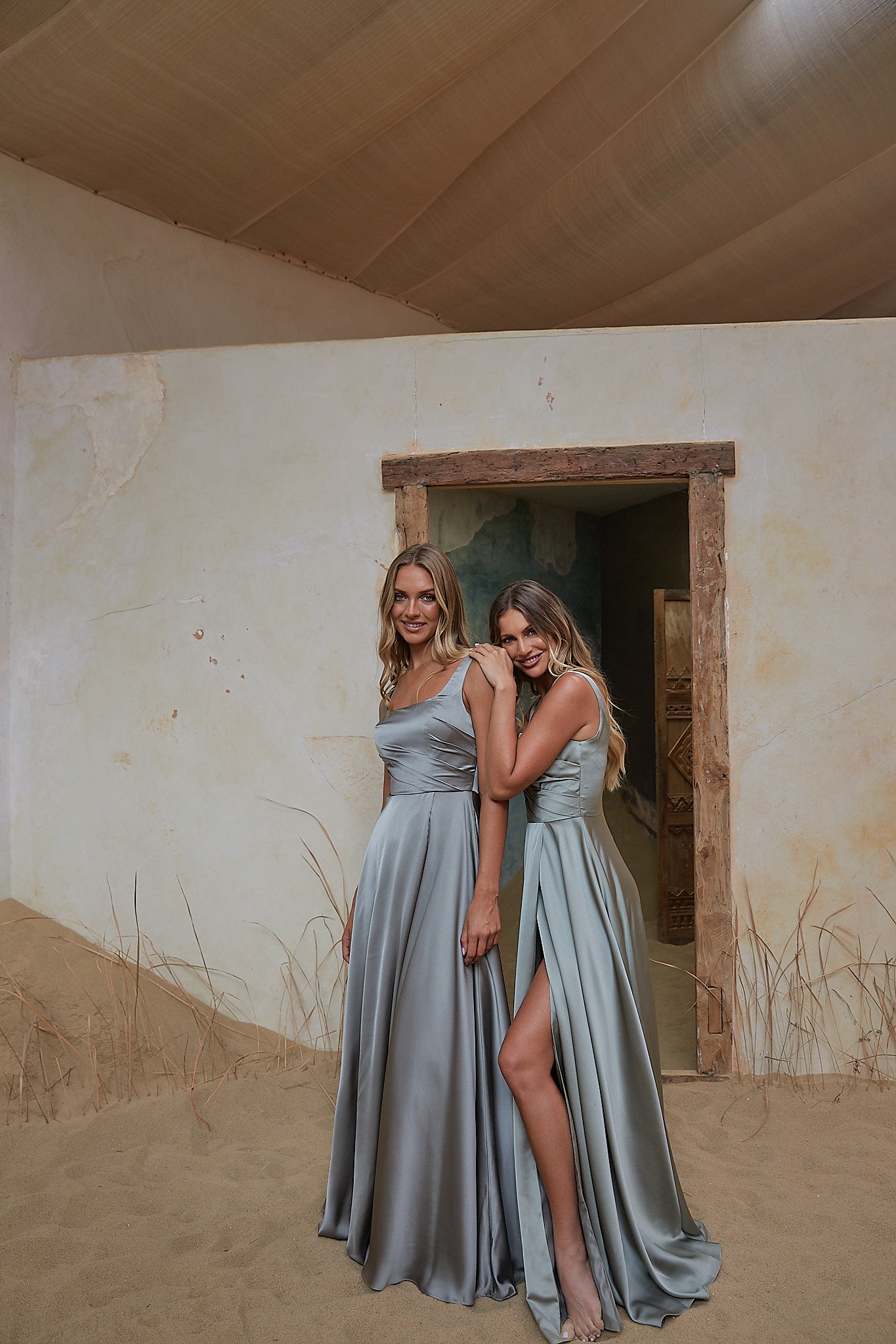 Two women wearing matching floor-length sage green satin bridesmaid dresses pose together in front of a rustic desert-style doorway with sandy walls and floor.