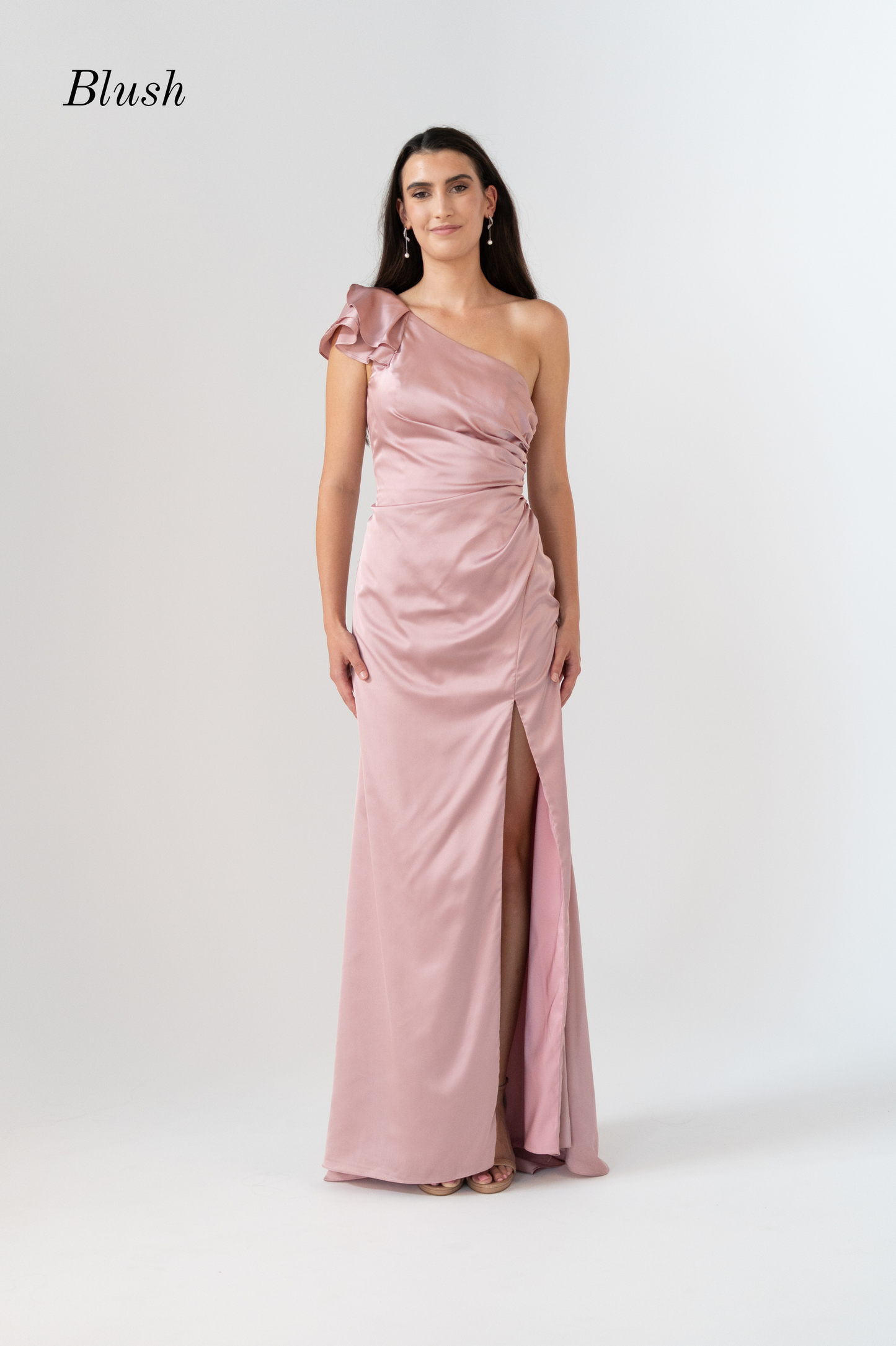 A woman modeling a blush pink one-shoulder floor-length gown with a gathered waist and thigh-high slit, photographed against a light gray studio background. The word 'Blush' appears in the upper left corner.