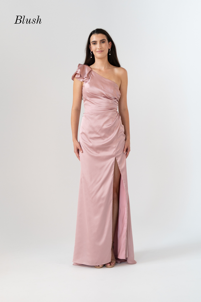 A woman modeling a blush pink one-shoulder floor-length gown with a gathered waist and thigh-high slit, photographed against a light gray studio background. The word 'Blush' appears in the upper left corner.
