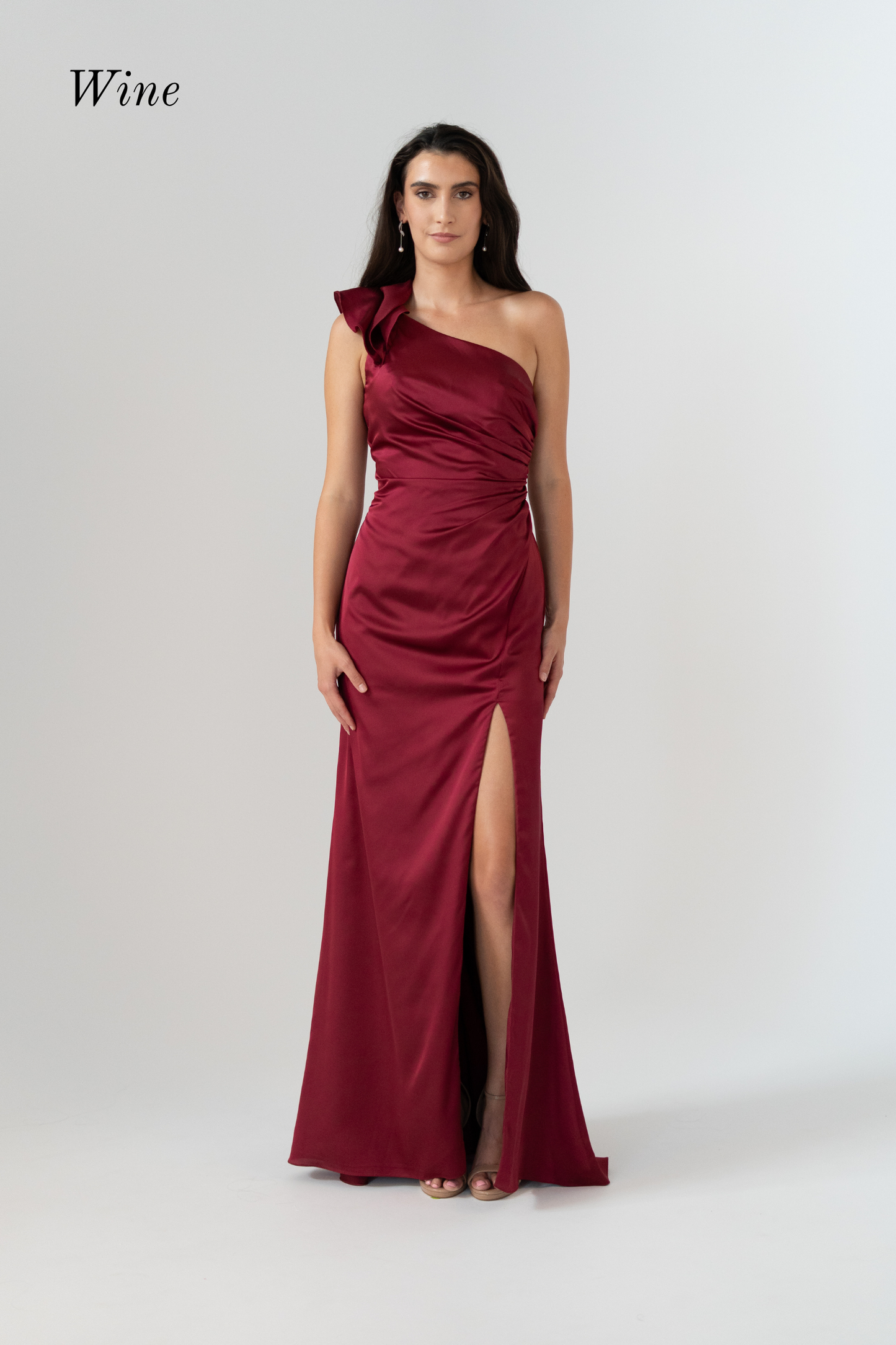 A woman modeling a wine-red satin one-shoulder evening gown with a high thigh slit, photographed against a light gray studio background. The word 'Wine' appears in the upper left corner.