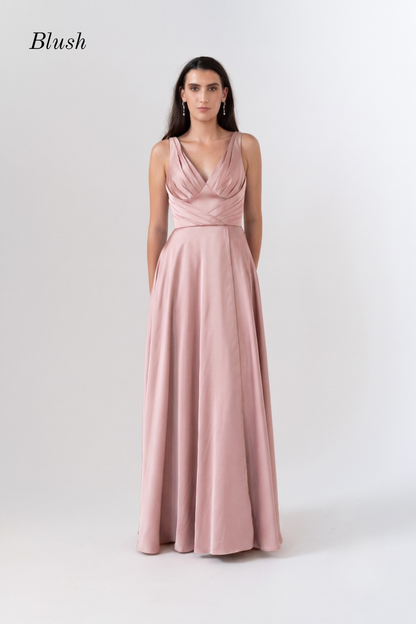 A woman modeling a blush pink sleeveless maxi dress with a draped, wrap-style bodice and a flowing skirt, shown against a light gray studio background with the brand name 'Blush' in the top left corner.