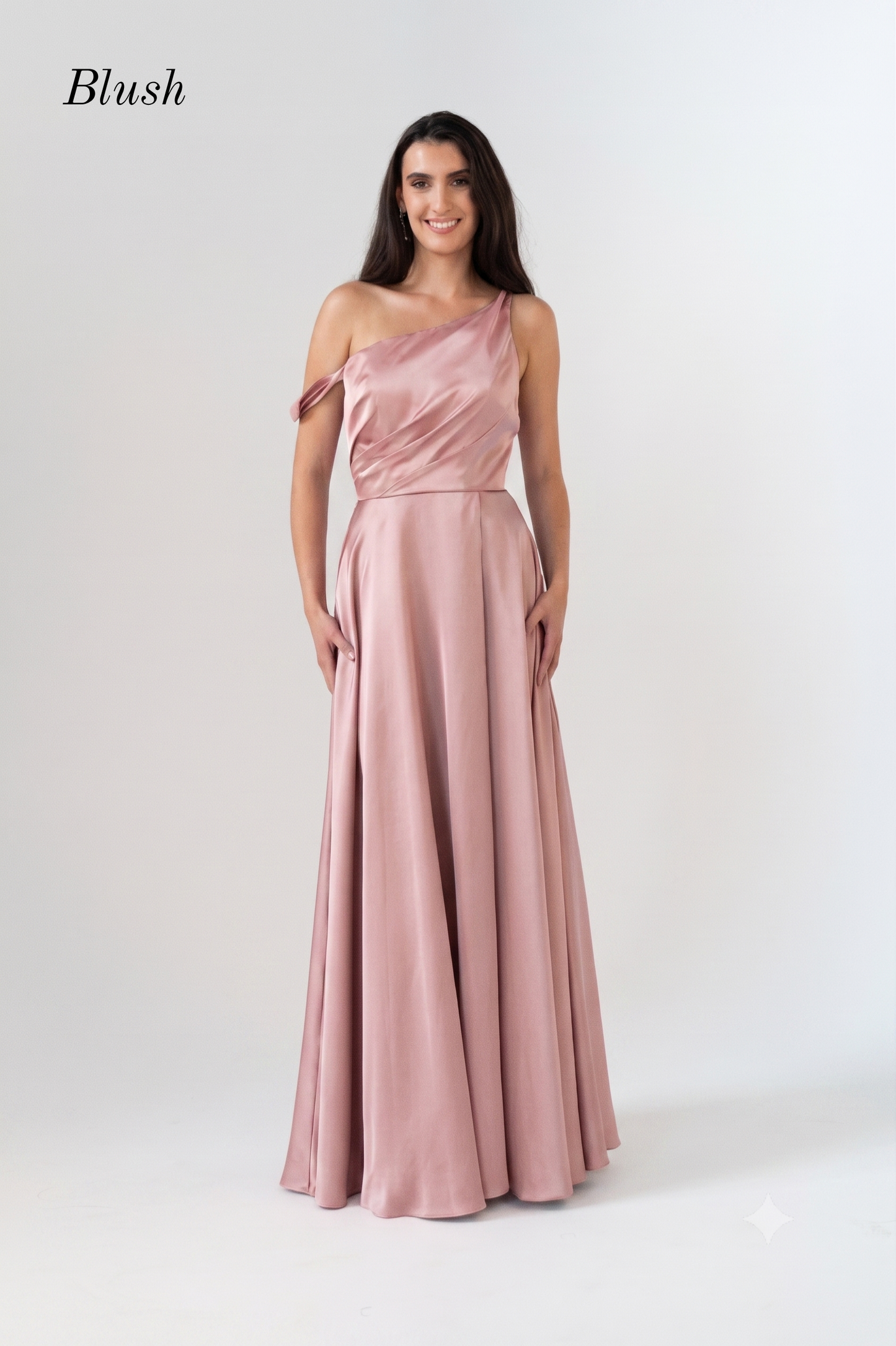 A woman modeling a blush pink one-shoulder floor-length gown with a draped bodice and flowing skirt, photographed against a plain light gray studio background. The word 'Blush' appears in the upper left corner.