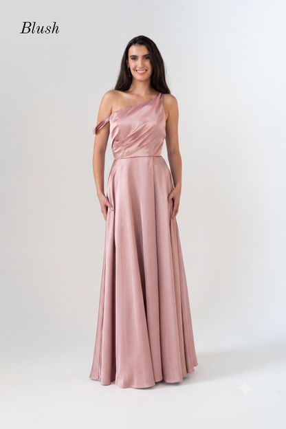 A woman modeling a blush pink one-shoulder floor-length gown with a draped bodice and flowing skirt, photographed against a plain light gray studio background. The word 'Blush' appears in the upper left corner.
