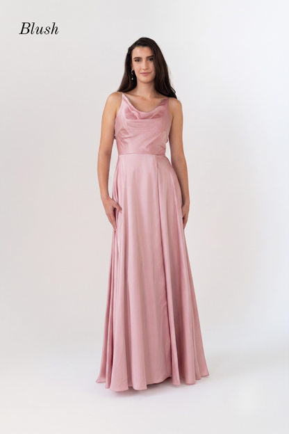 A woman modeling a blush pink floor-length gown with a draped neckline and sleeveless design, photographed against a plain light background. The word 'Blush' appears in the upper left corner.
