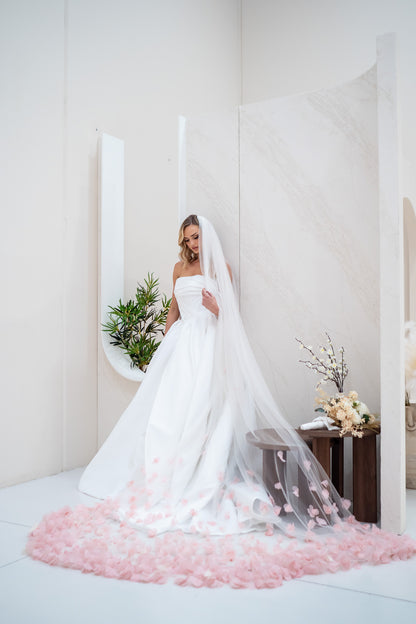 Bride in a white wedding dress with a long veil standing on pink petals in a minimalistic room.
