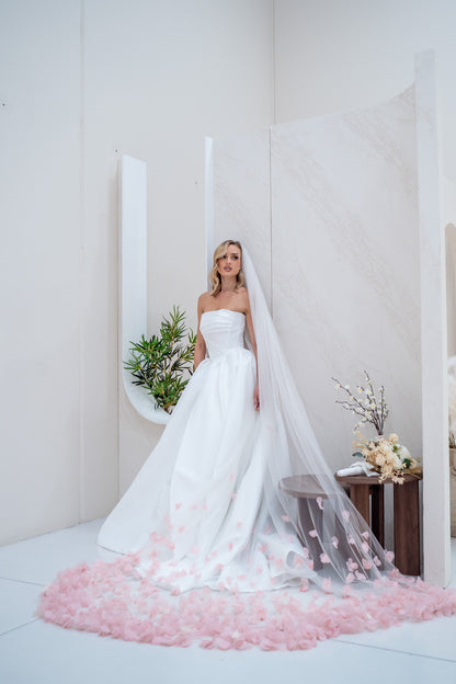 Bride in a strapless wedding dress with a long veil and pink petal skirt standing in a minimalistic room.