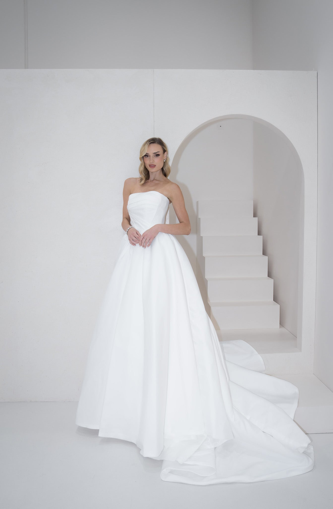 Woman in a white strapless gown standing in a minimalistic setting with a staircase.