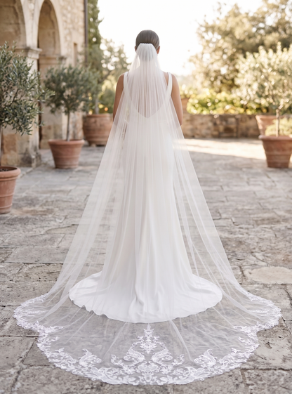Person wearing a long white wedding veil in an outdoor setting with stone pavement and greenery.