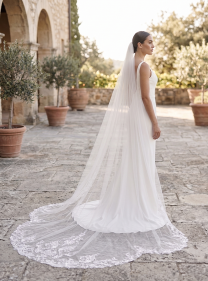 Woman in a white wedding dress with a long veil standing in an outdoor setting.