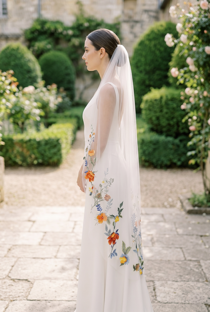 Woman in a floral embroidered dress and veil standing in a garden.