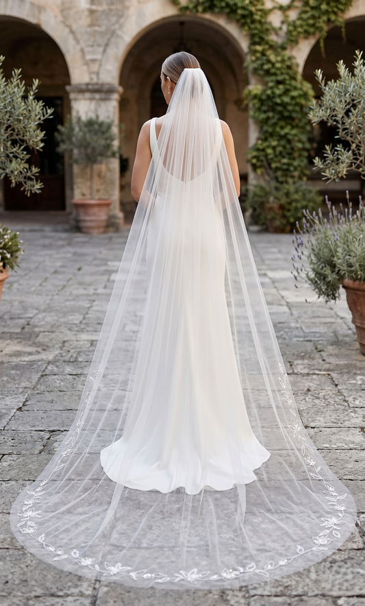 Bride wearing a long white veil in an outdoor setting with arches and plants.