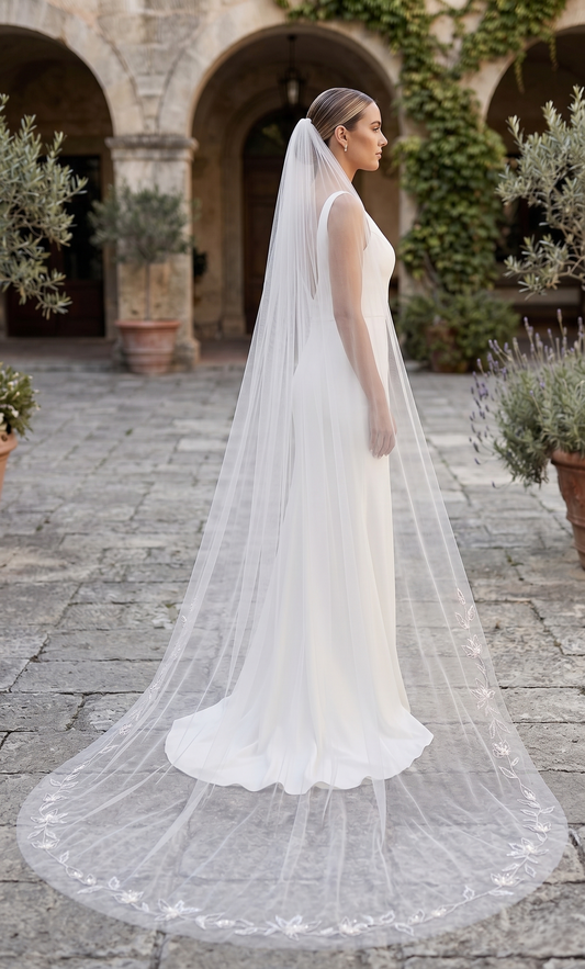 Bride in a white wedding dress with a long veil standing in an outdoor setting with arches and plants.