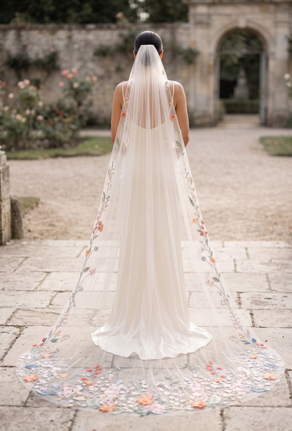 Bride with a long, floral-adorned veil standing in a garden setting.