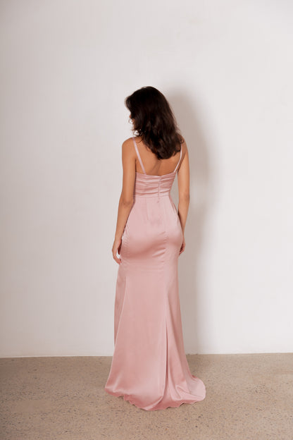 A woman stands with her back to the camera wearing a floor-length dusty pink satin gown with thin spaghetti straps and a ruched back detail, set against a plain off-white wall and light beige floor.
