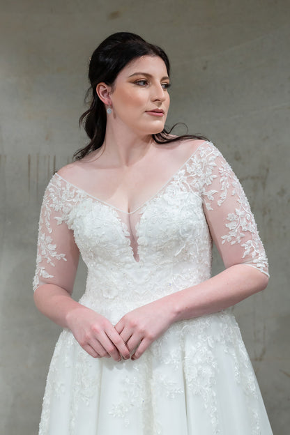 A woman wearing a white lace wedding dress with sheer long sleeves and a deep V-neckline, standing against a textured gray wall.